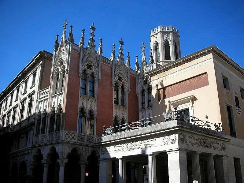 Facciata di edificio in stile gotico con guglie appuntite, finestre ad arco e una torre contro un cielo azzurro, affiancato da un edificio classico, tonalità calde di mattone e pietra