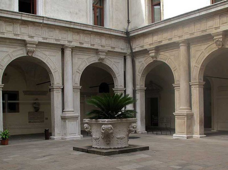 Cortile di un chiostro in pietra con colonnati ad arco, un pozzo scolpito in pietra al centro sormontato da una palma in vaso, tonalità neutre e calme