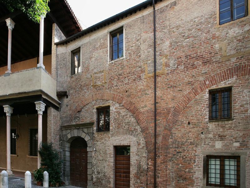 Esterno di un antico edificio in mattoni con portali ad arco, piccole finestre e una loggia coperta sorretta da colonne in pietra, tonalità terrose