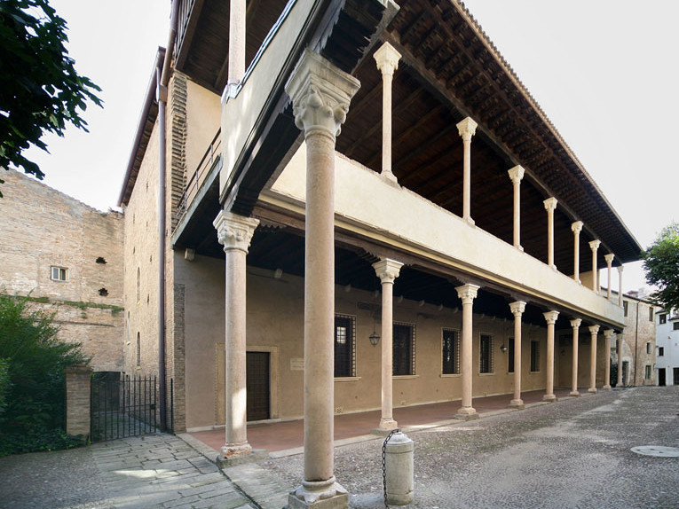 Edificio storico a due piani con loggia superiore in legno sostenuta da colonne in pietra, tonalità beige, cortile vuoto e silenzioso