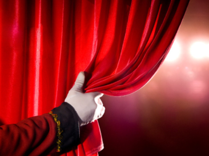 Primo piano di una mano guantata che scosta un pesante sipario di velluto rosso, con luci di scena che filtrano sullo sfondo, toni rossi e dorati